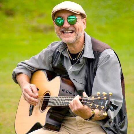 Older man wearing a beige cap and green sunglasses plays an acoustic guitar outdoors, smiling warmly.