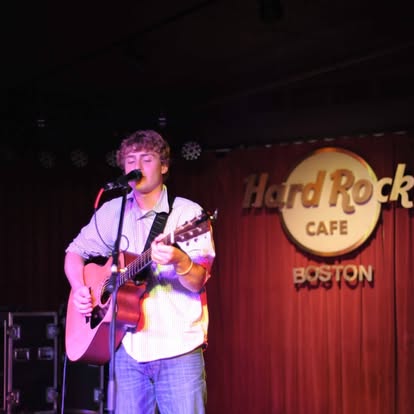 Male singer-guitarist performing on a stage at Hard Rock Cafe Boston, singing into a microphone with a pink-purple-lit backdrop.