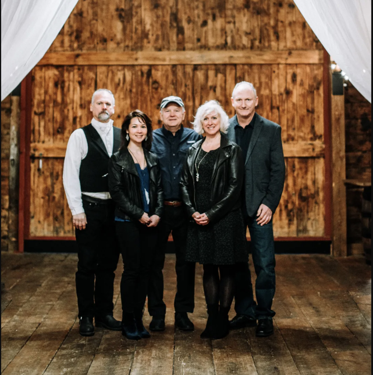 Five adults posing together in a rustic wooden room, two women in leather jackets and a man in a cap, all smiling at the camera.