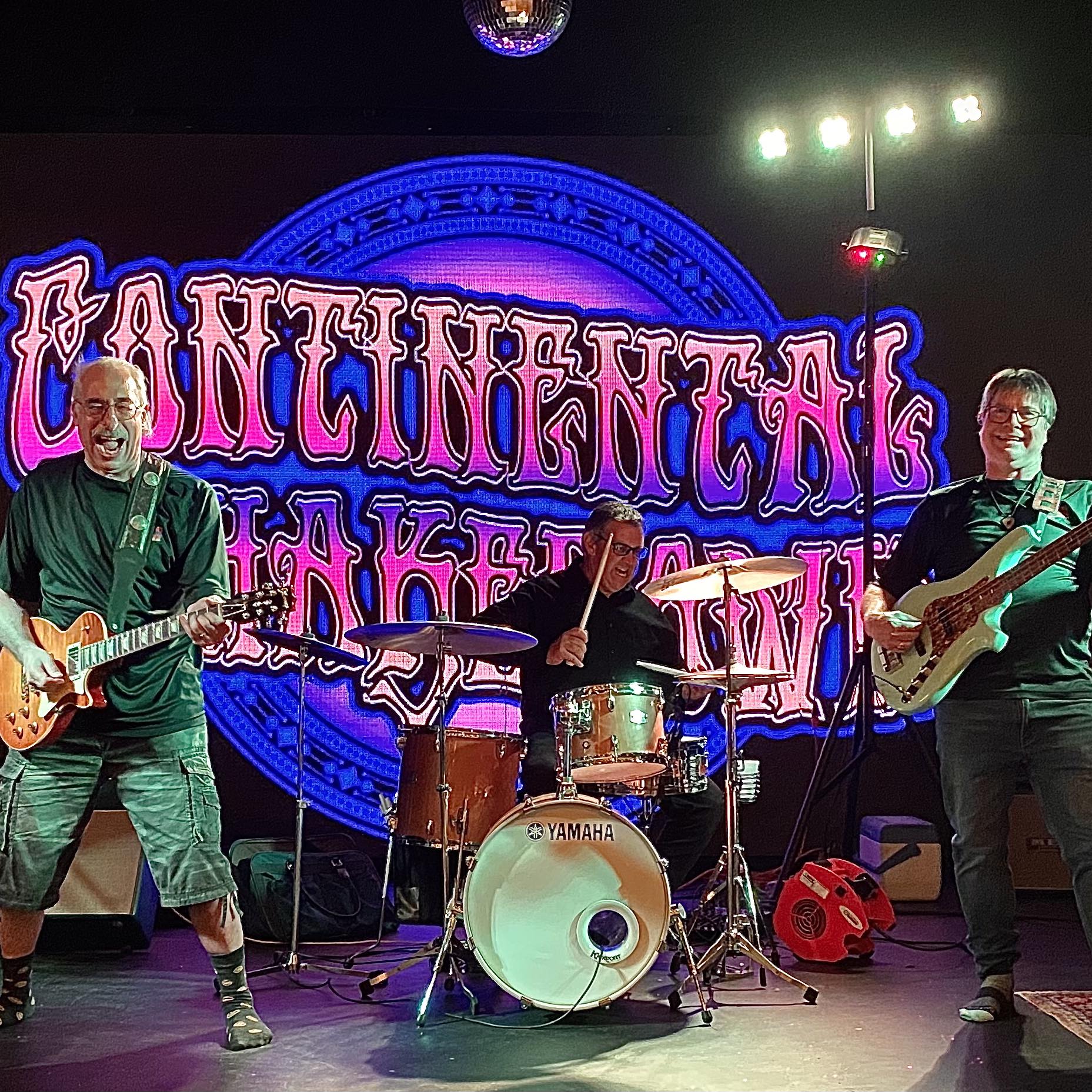 Guitarist left, drummer center, and bassist right performing on a stage with a bright neon blue backdrop and colorful lights behind them.