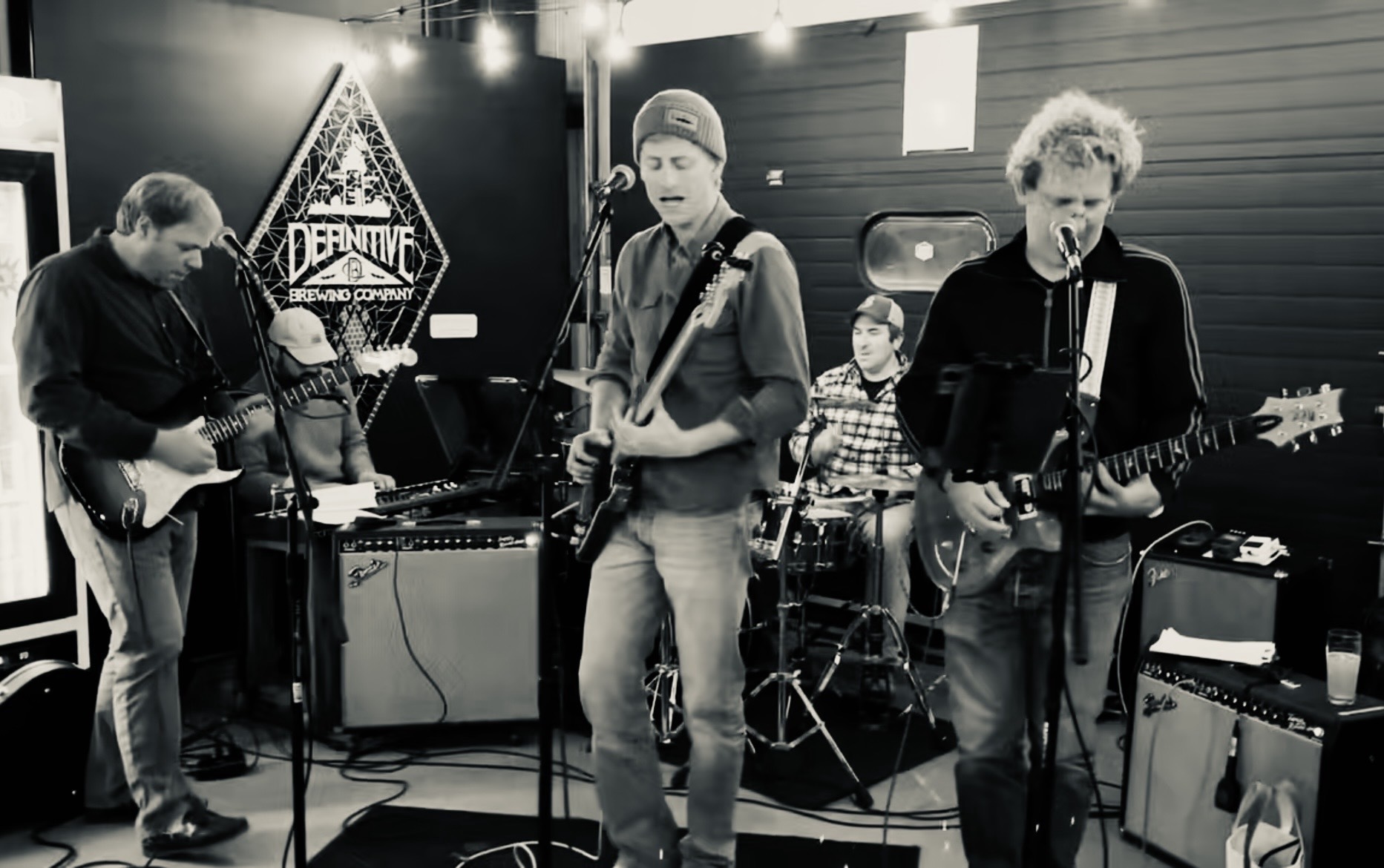 A four-member band performs in a brewery setting; two guitarists sing into microphones while a drummer plays in the back, with an amplifier and Definite Brewing Company signage visible in the background.
