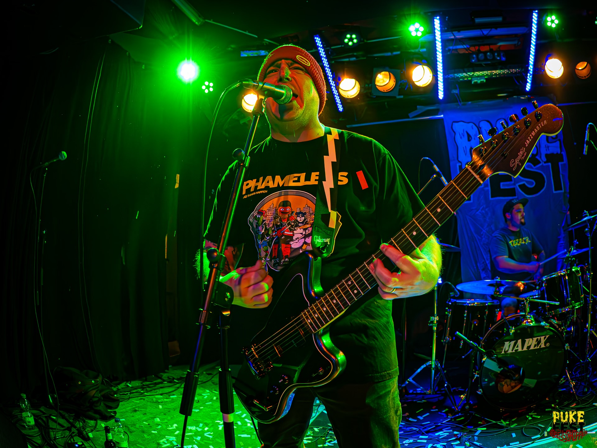 Guitarist wearing a beanie sings into a mic on a green-lit stage, playing a black electric guitar with a graphic T‑shirt visible.