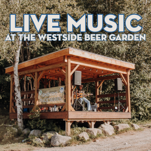 Open wooden stage in a wooded beer garden with two speakers; banner above reads 'LIVE MUSIC AT THE WESTSIDE BEER GARDEN'.