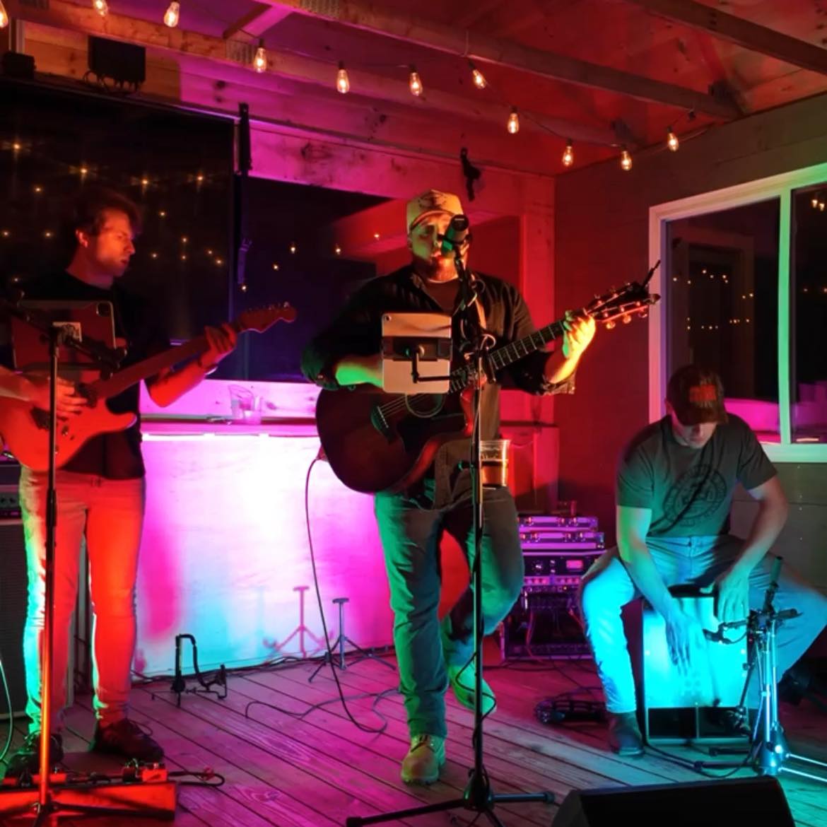 Three musicians perform on a small stage under colorful pink, purple, and blue lights: center singer with acoustic guitar, left guitarist, right percussionist seated with a cajón.