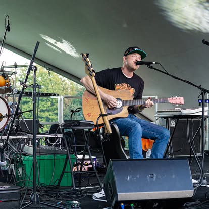 Musician in a black tee and cap playing an acoustic-electric guitar and singing into a microphone on an outdoor stage.