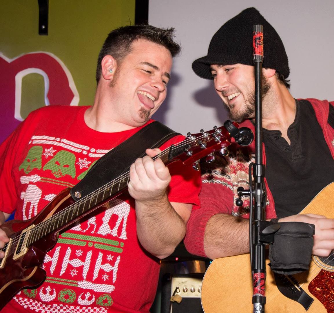 Two male musicians perform together on stage, smiling while playing guitars; one wears a red festive sweater and the other a black beanie, with a microphone between them.