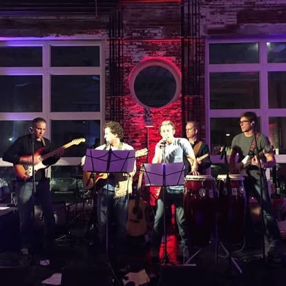 Five-piece band performing on a dimly lit stage with purple and red lights; guitarists on both ends, a vocalist center, and percussion with congas.