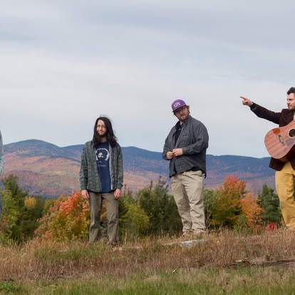 Four people outdoors on a grassy hill with autumn trees; a guitarist on the right and others listening as one points toward the distance.