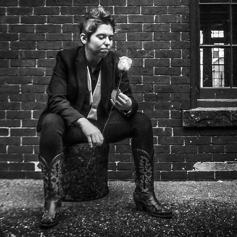 Person in a blazer and boots sits on a concrete block against a brick wall, holding a single rose and looking at it pensively, in black and white.
